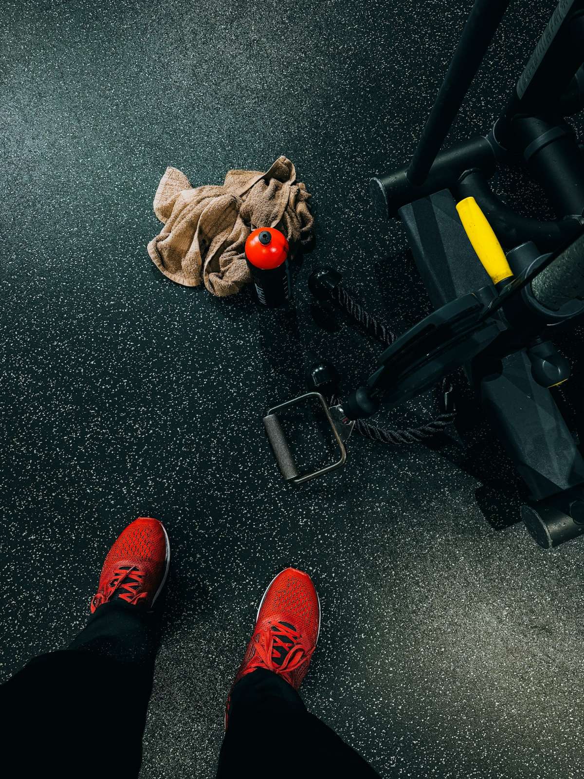 Overhead view of red training shoes on a speckled gym floor beside a towel, bottle and cable machine
